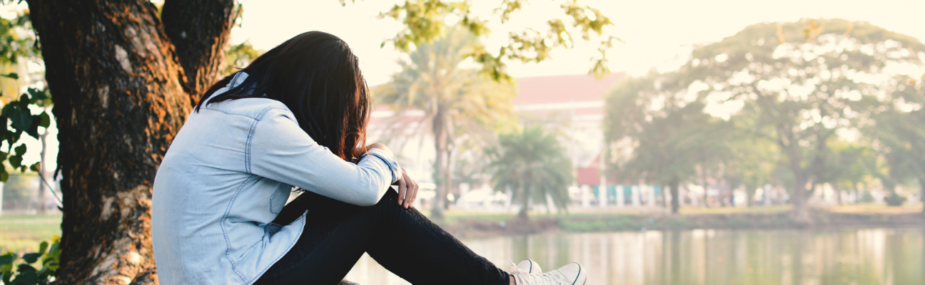 Person sitting by tree with head down and lake in background